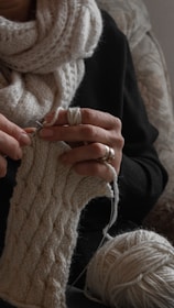 A person is knitting with beige yarn, creating a textured, patterned cloth. They are wearing a cozy scarf and a dark sweater. The focus is on their hands, which hold knitting needles and the partially completed knitting. A ball of yarn and their rings are also visible.
