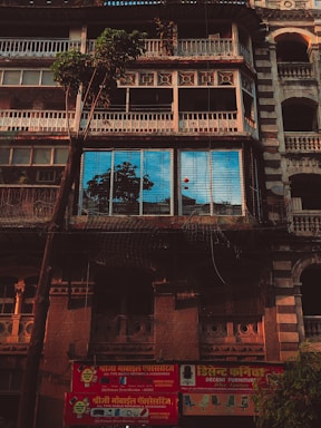 An old building with intricate architectural details, showcasing a facade with multiple balconies and a large tree in front. The windows reflect a bright blue sky and nearby structures. A red signboard in a local script advertises mobile repair and furniture services at the bottom of the building.