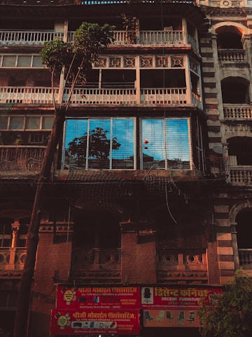 An old building with intricate architectural details, showcasing a facade with multiple balconies and a large tree in front. The windows reflect a bright blue sky and nearby structures. A red signboard in a local script advertises mobile repair and furniture services at the bottom of the building.