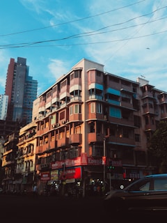 A bustling urban street scene features a multi-story building with various shops on the ground floor. The architecture exhibits a mix of modern and slightly older styles, with a combination of glass balconies and wooden shutters. Cars and people are visible, indicating a lively atmosphere. The sky is bright with scattered clouds, and there are electrical wires crisscrossing above.