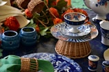 A vibrant tea set with ornate floral patterns in blue and orange sits arranged on a woven basket stand. Surrounding the set are decorative blue ceramic candle holders with heart cutouts, orange flowers, and various tableware pieces. The table is set on a deep blue surface accented with green napkins.