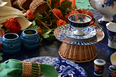A vibrant tea set with ornate floral patterns in blue and orange sits arranged on a woven basket stand. Surrounding the set are decorative blue ceramic candle holders with heart cutouts, orange flowers, and various tableware pieces. The table is set on a deep blue surface accented with green napkins.