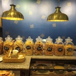 Several glass jars filled with various snacks are aligned on a granite counter. Labels with prices and names of the snacks are visible on each jar. Two large brass pendant lights hang above, illuminating the setup. The background features a blue wall decorated with a simple gold pattern.