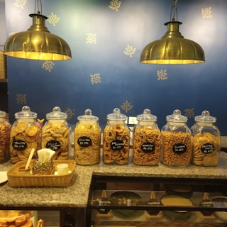 Several glass jars filled with various snacks are aligned on a granite counter. Labels with prices and names of the snacks are visible on each jar. Two large brass pendant lights hang above, illuminating the setup. The background features a blue wall decorated with a simple gold pattern.