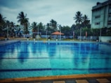 A large swimming pool with clear blue water is surrounded by a tiled deck. In the background, there are tall palm trees and a few buildings, providing a sense of a tropical setting. A small pavilion with a red roof can be seen near the pool area, adding a touch of leisure and relaxation.