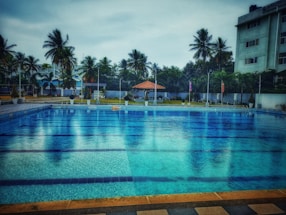 A large swimming pool with clear blue water is surrounded by a tiled deck. In the background, there are tall palm trees and a few buildings, providing a sense of a tropical setting. A small pavilion with a red roof can be seen near the pool area, adding a touch of leisure and relaxation.