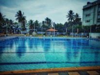A large swimming pool with clear blue water is surrounded by a tiled deck. In the background, there are tall palm trees and a few buildings, providing a sense of a tropical setting. A small pavilion with a red roof can be seen near the pool area, adding a touch of leisure and relaxation.