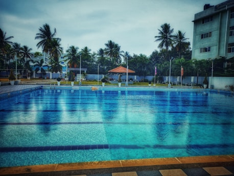A large swimming pool with clear blue water is surrounded by a tiled deck. In the background, there are tall palm trees and a few buildings, providing a sense of a tropical setting. A small pavilion with a red roof can be seen near the pool area, adding a touch of leisure and relaxation.