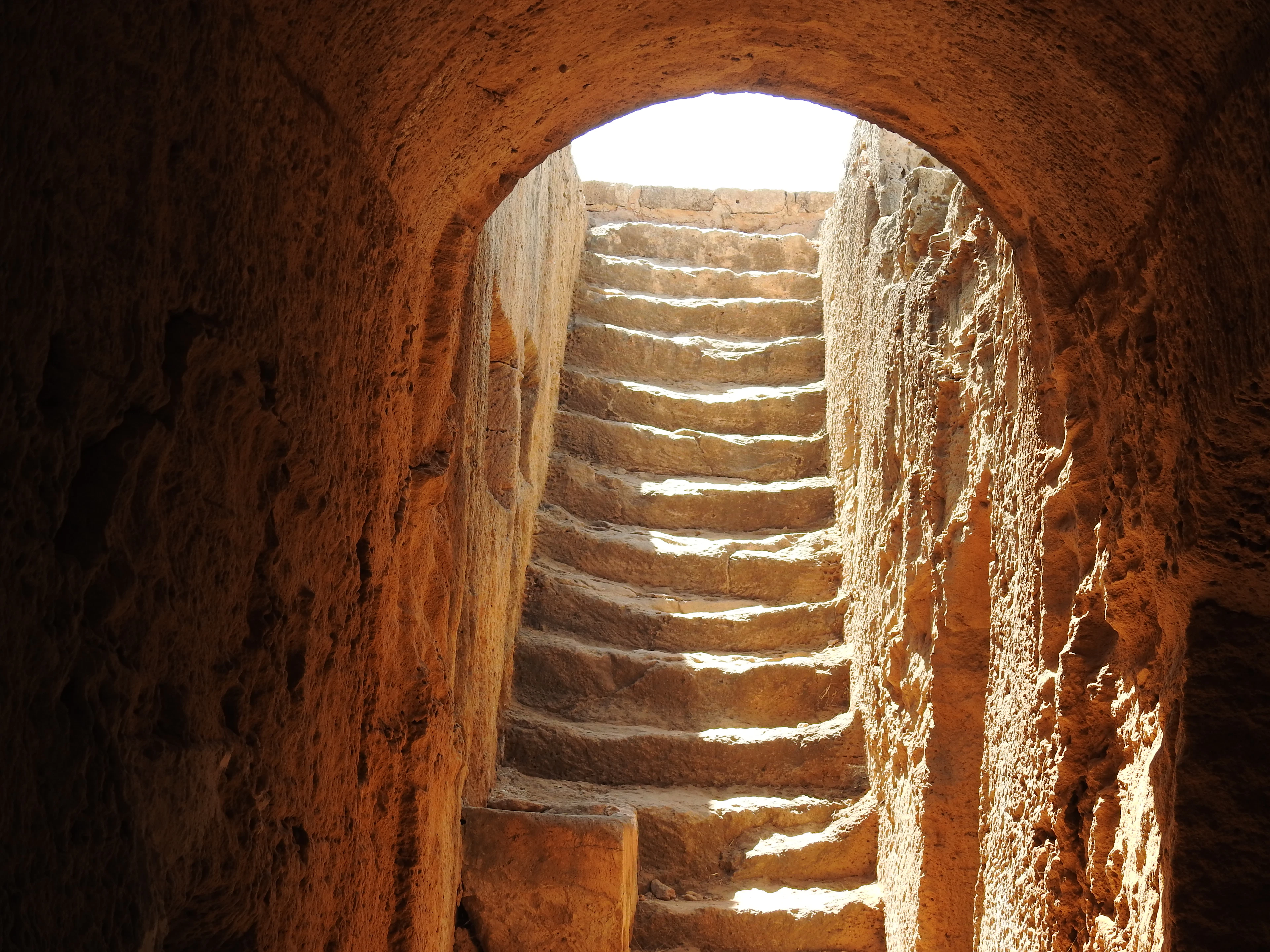 A rock-cut stairway descending into an underground tomb at the Tombs of the Kings in Paphos, its honey-coloured limestone walls glowing where daylight spills down the worn steps from the opening above