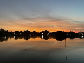 The image features a serene lakeside scene at sunset with vibrant orange and blue colors in the sky. The water reflects the sky and nearby trees, creating a mirror-like effect. Silhouettes of trees line the horizon, and small pinpoints of light suggest distant buildings or streetlights.