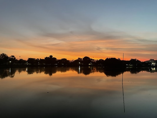 The image features a serene lakeside scene at sunset with vibrant orange and blue colors in the sky. The water reflects the sky and nearby trees, creating a mirror-like effect. Silhouettes of trees line the horizon, and small pinpoints of light suggest distant buildings or streetlights.