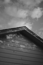 A monochrome image depicting the gable end of a house roof. Shadows of nearby trees are visible on the siding, creating a textured pattern. The sky is partly cloudy, adding depth to the background.