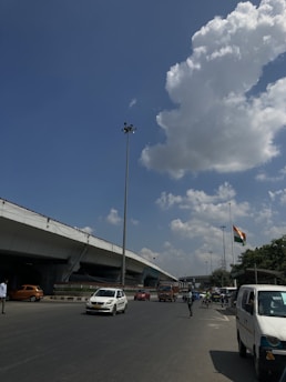 A busy urban street scene features several vehicles including cars, a truck, and rickshaws on the road. A large overpass runs parallel to the street. The national flag is visible waving in the distance. The sky is mostly clear with some large white clouds.