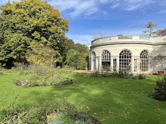 A well-maintained garden with lush green grass and vibrant flowers, featuring a classical white building with large arched windows in the background. A large, leafy tree provides shade at the corner of the garden.