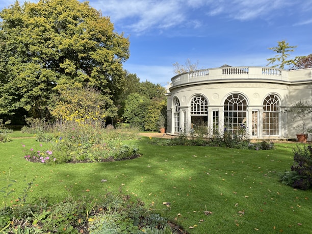 A well-maintained garden with lush green grass and vibrant flowers, featuring a classical white building with large arched windows in the background. A large, leafy tree provides shade at the corner of the garden.