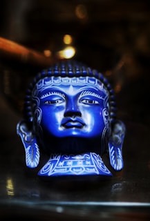 A blue ceramic sculpture of a head, intricately designed with white patterns and detailed facial features, including closed eyes and serene expression. The background is softly blurred, adding a glowing effect to the sculpture.