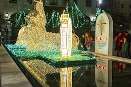 A festive light display showcases a large illuminated sculpture reflecting in a pool of water. The structure includes intricate wireframe designs lit with golden and green lights. Nearby, people observe the display, and a monument marker reads 'Año Jubilar Lebaniego 2023.' Trees adorned with green lights add to the ambiance.