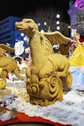 A sand sculpture of a dragon with intricate details is displayed in an outdoor setting at night. Surrounding the sculpture are festive lights, decorations, and snow-like material on the ground. Several other sculptures and decorations, including a figure in a yellow dress, can be seen in the background.
