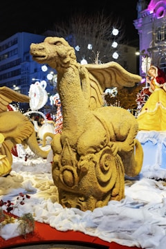 A sand sculpture of a dragon with intricate details is displayed in an outdoor setting at night. Surrounding the sculpture are festive lights, decorations, and snow-like material on the ground. Several other sculptures and decorations, including a figure in a yellow dress, can be seen in the background.