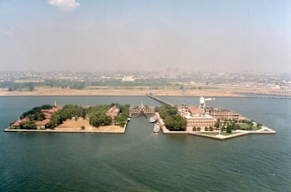 Aerial view of two small, lush green islands surrounded by water. The island on the right has a distinctive building complex with red-roofed structures, while the left island has smaller buildings scattered among trees. Several boats are docked between the islands, and a hazy city skyline is visible in the background.