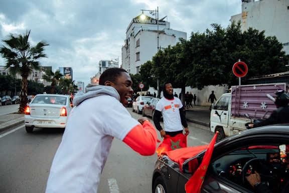 Two people on a lively street are interacting joyfully. One of them is leaning towards a car with an open window and a Moroccan flag draped over it, while the other stands holding the flag. Cars are stopped on a busy road lined with trees and a hotel sign visible in the distance. The atmosphere appears festive with various pedestrians and vehicles surrounding the scene.