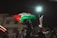 A person stands holding a large flag with red, black, white, and green sections, associated with Palestine. The scene is set at night, illuminated by streetlights. Surrounding the person, several other people appear, some partially visible. Buildings and street decorations with lights are in the background.