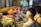 A vibrant market scene with fresh produce such as tomatoes, gourds, cilantro, and pineapples displayed prominently. Two individuals engage in a transaction, exchanging packaged goods. The setting is bustling, indicative of a lively marketplace.