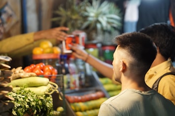 A vibrant market scene with fresh produce such as tomatoes, gourds, cilantro, and pineapples displayed prominently. Two individuals engage in a transaction, exchanging packaged goods. The setting is bustling, indicative of a lively marketplace.