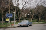 A vintage blue car is parked on a quiet street beside a sign that reads 'Otopark'. The scene is set in a leafy area with bare trees and a fence lining the background. The atmosphere is tranquil, reflecting an overcast day.
