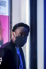 A person wearing a black face mask and dark clothing is positioned in a dimly lit setting, looking directly at the camera. The background includes a brightly lit display with a pink and white panel, possibly a poster or advertisement, partially visible on the left side.