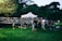 Photo of a white event tent set up in a sunny park with people gathering underneath.