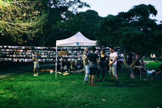 A group of people gathers around a white tent in a park setting, with numerous photographs displayed on strings. The environment is lush with grass and trees, and it appears to be an outdoor photo exhibition or community event.