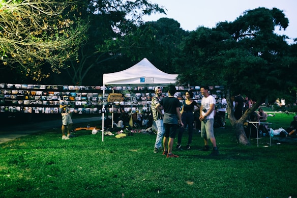 A group of people gathers around a white tent in a park setting, with numerous photographs displayed on strings. The environment is lush with grass and trees, and it appears to be an outdoor photo exhibition or community event.