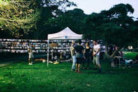 A group of people gathers around a white tent in a park setting, with numerous photographs displayed on strings. The environment is lush with grass and trees, and it appears to be an outdoor photo exhibition or community event.