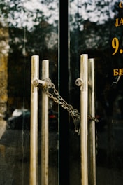 A pair of glass doors with vertical metal handles are secured by a metal chain. The reflective surface of the doors shows a faint reflection of an outdoor scene, including trees and a building. The handles are cylindrical and aligned parallel to each other.