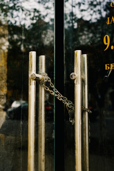 A pair of glass doors with vertical metal handles are secured by a metal chain. The reflective surface of the doors shows a faint reflection of an outdoor scene, including trees and a building. The handles are cylindrical and aligned parallel to each other.