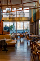 A stylish and modern restaurant interior with wooden accents and ample seating. Large windows provide a view of the cityscape outside, allowing natural light to fill the space. Hanging lights with a unique design add to the contemporary ambiance. Tables are set with dishes and glassware, ready for guests.