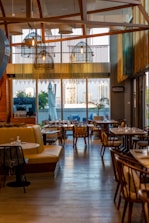 A stylish and modern restaurant interior with wooden accents and ample seating. Large windows provide a view of the cityscape outside, allowing natural light to fill the space. Hanging lights with a unique design add to the contemporary ambiance. Tables are set with dishes and glassware, ready for guests.