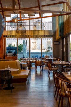 A stylish and modern restaurant interior with wooden accents and ample seating. Large windows provide a view of the cityscape outside, allowing natural light to fill the space. Hanging lights with a unique design add to the contemporary ambiance. Tables are set with dishes and glassware, ready for guests.