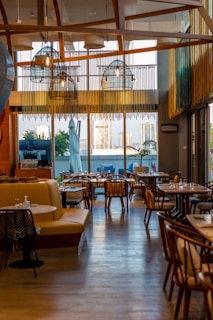 A stylish and modern restaurant interior with wooden accents and ample seating. Large windows provide a view of the cityscape outside, allowing natural light to fill the space. Hanging lights with a unique design add to the contemporary ambiance. Tables are set with dishes and glassware, ready for guests.