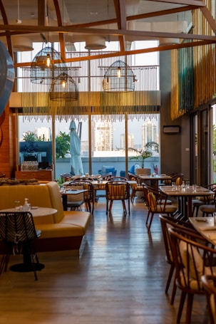 A stylish and modern restaurant interior with wooden accents and ample seating. Large windows provide a view of the cityscape outside, allowing natural light to fill the space. Hanging lights with a unique design add to the contemporary ambiance. Tables are set with dishes and glassware, ready for guests.