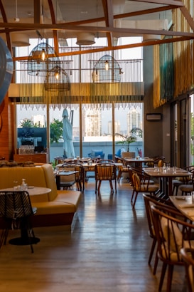 A stylish and modern restaurant interior with wooden accents and ample seating. Large windows provide a view of the cityscape outside, allowing natural light to fill the space. Hanging lights with a unique design add to the contemporary ambiance. Tables are set with dishes and glassware, ready for guests.