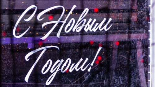 Stylish white script text in Cyrillic is overlaid on a background with a dark, reflective surface. Small, out-of-focus red and orange lights appear in the background, suggesting festivity or a celebration.