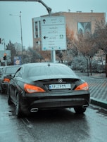 A black Mercedes car is stopped on a wet road with its taillights illuminated. Road signs in both Persian and English are visible, providing directions to various locations. The surroundings include trees and a building with decorative artwork.