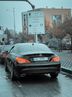 A black Mercedes car is stopped on a wet road with its taillights illuminated. Road signs in both Persian and English are visible, providing directions to various locations. The surroundings include trees and a building with decorative artwork.