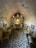 A small, rustic room with stone walls and a cobbled floor. There are several simple, wooden chairs arranged along the walls. In the center of the back wall, there is a small altar with flower arrangements forming the word 'DAD'. A narrow window is situated above the altar, illuminating the area with natural light. Various items, including framed pictures and a corkboard, adorn the walls.