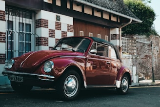 A classic red Volkswagen Beetle is parked on a street beside a building with a patterned facade featuring both brick and white square accents. The car's black convertible roof is up, and its shiny chrome details reflect the sunlight. Nearby, the building's windows have decorative metal bars, and there is greenery visible above the structure.