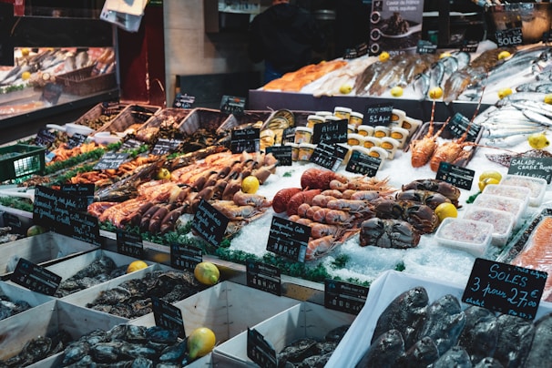A bustling seafood market display with a wide array of fresh seafood including fish, crabs, lobsters, oysters, and shrimp. The seafood is arranged on a bed of ice with signs indicating prices and species. Vibrant lemons are scattered throughout, adding a touch of color. The market stall is well-stocked and organized, conveying a sense of abundance and freshness.
