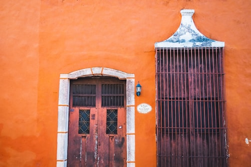 An orange stucco wall with a weathered wooden door and a large barred window. The door features stone arches and small square windows with wrought iron details. A black lantern-style light fixture is mounted next to the door, and a small oval plaque with text is affixed nearby. The wall's vivid color contrasts with the aged textures of the door and window.