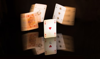 A collection of playing cards, with an Ace of Hearts prominently displayed in the foreground. The cards are arranged in such a way that they appear to be floating or suspended, and their soft reflections are visible on a glossy black surface. The image is creatively blurred in certain areas, giving a dynamic and energetic feeling to the composition.